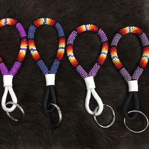 Op de afbeelding: Vier sleutelhangers met kralen en een lusontwerp. Elke sleutelhanger heeft een ander kleurenschema: paars, blauw, oranje en rood. De kralen lussen zijn bevestigd aan een wit of zwart koord met een zilveren sleutelring.