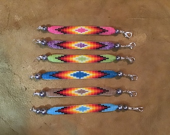 Könnte beinhalten: Sechs Perlenarmbänder mit geometrischem Muster in verschiedenen Farben, darunter Pink, Lila, Grün, Blau, Braun und Hellblau. Jedes Armband hat einen silbernen Verschluss.