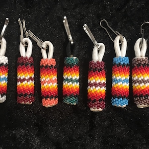Op de afbeelding: Een verzameling van zeven kralen rits trekt met een verscheidenheid aan kleurrijke patronen. Elke trekker heeft een cilindrisch kralenontwerp in tinten rood, oranje, geel, blauw en wit, bevestigd aan een wit koord en een zilverkleurige sluiting. De kralen secties zijn ongeveer 2,5 cm lang.