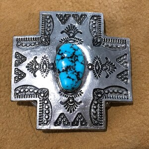 May include: A silver-colored cross-shaped pendant with intricate embossed patterns. The center features a vibrant, oval-shaped turquoise stone with black veining. The pendant has a rustic, Southwestern aesthetic.