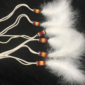 May include: A collection of white feather pendants with beaded details. Each pendant has a bunch of fluffy white feathers connected to a beaded cylinder with colourful patterns of red, orange, yellow, and blue. White leather cords extend from the beaded sections.