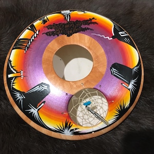 Pode incluir: Um tambor de madeira com um desenho pintado de um pôr do sol e um apanhador de sonhos. O tambor é pintado com cores laranja, amarelo, roxo e preto. O apanhador de sonhos é branco com uma conta azul.