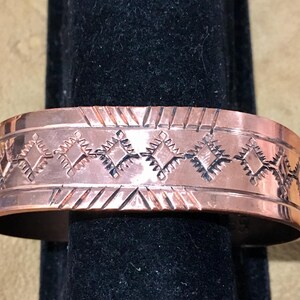 May include: A copper cuff bracelet with an etched geometric pattern. The bracelet has a polished finish and is displayed on a black stand. The design features diamond and line shapes, creating a decorative accessory.