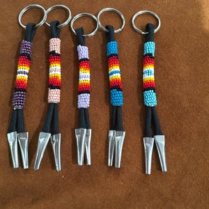 May include: Five beaded keychains with silver-colored metal clasps. Each keychain features a different color pattern of small beads, including red, orange, yellow, blue, and purple, strung on black cords.