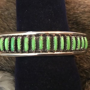 May include: A silver cuff bracelet featuring a single horizontal row of seventeen vertical, oval-shaped, bright green stones set in individual serrated metal bezels. The polished silver band has an open design with smooth, rounded edges.