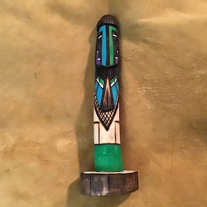 Puede incluir: Un tótem de madera con una cara tallada y patrones geométricos pintados en azul, verde, morado y blanco. El tótem está de pie sobre una base de madera.