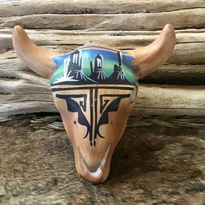 May include: A wooden bull skull with painted details. The skull features a landscape design in blue, green, and black. The horns are a light brown color. The skull is resting on a textured surface.