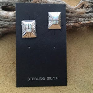 Puede incluir: Un par de pendientes cuadrados de plata de ley. Cada pendiente presenta una piedra central similar al ópalo rodeada por un diseño plateado de rayos de sol. Los pendientes se exhiben en una tarjeta negra con "STERLING SILVER" impreso.