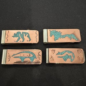 Può includere: Quattro fermasoldi in rame con disegni di animali intarsiati in turchese. Ogni fermasoldi ha un'estremità color argento. I disegni includono un orso, un cavallo e altri due animali. I fermasoldi sono rettangolari e lucidi.