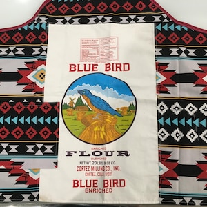 以下が含まれることがあります： 青い鳥のイラストと「Blue Bird Enriched Flour Bleached Net Wt. 20 lbs. 9.08 kg. Cortez Milling Co., Inc. Cortez, Colo. 81321 Blue Bird Enriched」というテキストが書かれた白い小麦粉袋。袋は、幾何学模様の黒と赤の模様の布地に付けられています。
