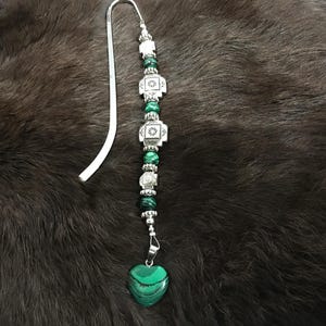 May include: A silver bookmark with a green heart-shaped charm and green and silver beads. The bookmark is on a brown furry surface.