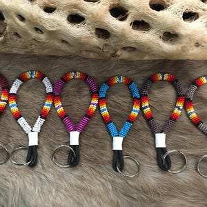 May include: Six beaded wrist lanyards in various colors, including red, white, purple, blue, brown, and gray. Each lanyard features a loop of colorful beads, a white base, a black cord, and a silver keyring. The lanyards are displayed on a furry surface.