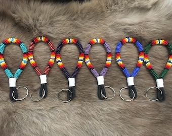 May include: Six beaded keychains in various colors, including turquoise, red, purple, blue, and green. Each keychain features a loop of colorful beads, a white band, a black cord, and a silver key ring. The keychains are displayed on a furry surface.
