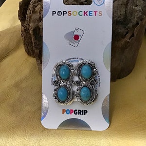 May include: A PopSockets PopGrip with four silver-toned, floral-designed grips, each featuring a turquoise-colored stone. The PopGrip is on a white and silver backing with the PopSockets and PopGrip logos.