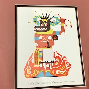 Puede incluir: Una ilustración colorida de un Kachina Ogre Negro, una figura tradicional de espíritu Hopi, con un cuerpo con patrones rojos, amarillos y blancos, un tocado azul y amarillo, y una mano roja sosteniendo un arco y una flecha. La figura se encuentra sobre una base naranja fuego.