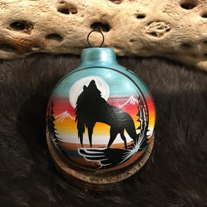 May include: A hand-painted glass ornament featuring a silhouette of a wolf howling at the moon. The ornament has a blue, turquoise, and orange background with a white moon and a mountain range.