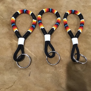 May include: Three beaded keychains with a black cord loop and silver key ring. The beaded portion features a pattern of black, white, red, orange, and blue beads. The keychains are displayed on a brown surface.