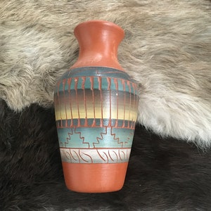 Könnte beinhalten: Eine kleine, terrakottafarbene Vase mit einem südwestlich inspirierten Design. Die Vase ist mit Bändern aus türkisfarbener, brauner, gelber und weißer Farbe verziert. Das Design umfasst geometrische Formen und stilisierte Blätter.