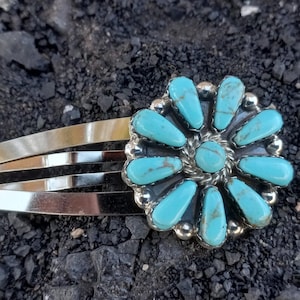 May include: A silver-toned metal hair clip featuring a circular floral-shaped ornament. The ornament is inlaid with ten elongated, teardrop-shaped turquoise stones surrounding a central round turquoise stone, all set in a polished silver frame.