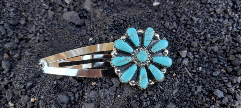 May include: A silver-toned metal hair clip featuring a circular floral-shaped ornament. The ornament is inlaid with ten elongated, teardrop-shaped turquoise stones surrounding a central round turquoise stone, all set in a polished silver frame.