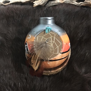 May include: Hand-painted ceramic dream catcher ornament. The ornament features a sunset scene with a dream catcher web, a turquoise stone, and a feather. The top is silver, and the bottom is orange and brown.