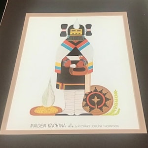 Puede incluir: Una ilustración colorida de una Maiden Kachina, una figura tradicional de espíritu Hopi, con un tocado blanco, ropa negra y blanca y un escudo rojo y negro. La obra de arte se titula "Maiden Kachina 1974 by Richard Joseph Thompson".