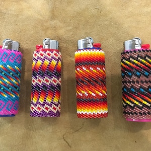 May include: Four lighters encased in intricate, hand-woven glass seed bead covers. Each cover features a unique geometric pattern in vibrant color combinations including turquoise, magenta, orange, yellow, red, black, and white glass beads.