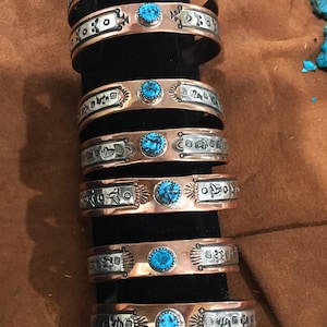 May include: A collection of copper and silver-tone cuff bracelets. Each bracelet features intricate etched designs and a central turquoise stone. The bracelets are displayed on a black stand against a brown background.