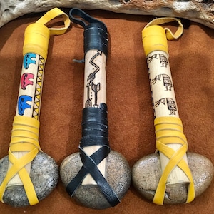 Puede incluir: Tres herramientas artesanales de piedra y madera. Los mangos están decorados con diseños coloridos de animales y geométricos, envueltos en cuero amarillo. Las cabezas de piedra están aseguradas con correas de cuero. Un mango es negro y dos son amarillos.