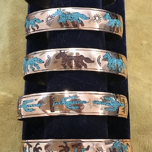 May include: Five copper cuff bracelets featuring inlaid turquoise and dark stone designs. The patterns include running horses, sun symbols, cacti, and stylized figures. Each bracelet has a polished metallic finish and a consistent band width.
