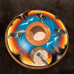 Pode incluir: Um tambor redondo de madeira com um design pintado em azul, laranja e amarelo. O tambor apresenta um apanhador de sonhos com uma pedra turquesa e uma pena. O tambor é decorado com beija-flores pintados.