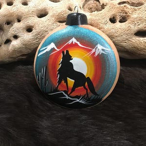 May include: A round, wooden Christmas ornament with a painted design of a coyote howling at the moon in a desert landscape. The background is a gradient of blue, orange, and yellow, with white mountains in the distance.
