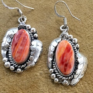 May include: A pair of silver dangle earrings featuring oval-shaped orange and red spiny oyster stones. The stones are set in polished silver frames decorated with leaf motifs, small silver beads, and twisted wire accents with hook backings.