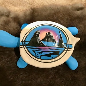 May include: A blue ceramic turtle with a white shell. The shell is painted with a black and white design of a mountain range and a lake. The design is surrounded by a black circle.