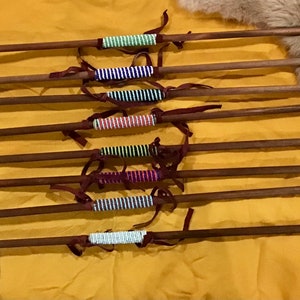 Puede incluir: Un conjunto de ocho flechas con plumas negras y envolturas de cuentas de colores. Las flechas están dispuestas sobre un fondo de tela amarilla.