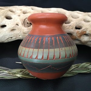 Puede incluir: Un pequeño jarrón de cerámica de terracota pintado a mano con un diseño geométrico en tonos marrones, verdes y azules. El jarrón tiene un cuerpo redondeado y un borde ligeramente acampanado.