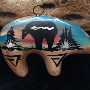 Op de afbeelding: Handbeschilderde houten berenornament. De zwarte beer staat voor een zonsondergang met bergen en bomen. Het ornament heeft een zwart touwtje om op te hangen en geometrische ontwerpen.