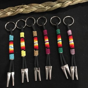 Op de afbeelding: Zes sleutelhangers met kralen en zilverkleurige metalen accenten. Elke sleutelhanger heeft een ander kleurenpatroon van kleine kralen, waaronder blauw, geel, rood, goud en paars, geregen op zwarte koorden. De sleutelhangers zijn gerangschikt op een zwart oppervlak.