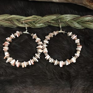 May include: A pair of large hoop earrings made of natural seashells. The earrings are a light pink and white color and have a silver hook closure.
