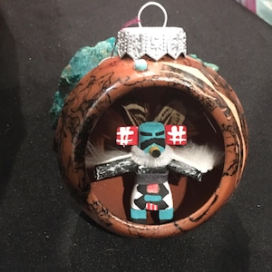 Puede incluir: Adorno navideño artesanal con una muñeca kachina nativa americana dentro de un adorno decorativo redondo marrón y negro. La muñeca está pintada con detalles azules, rojos, negros y blancos. El adorno tiene una tapa plateada y un lazo de metal para colgar.