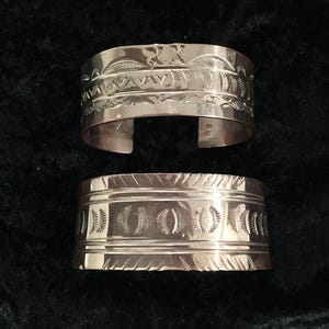 May include: Two silver-toned cuff bracelets with intricate etched designs. The top bracelet features a series of geometric and curved patterns. The bottom bracelet has a repeating pattern of oval shapes. Both are displayed on a dark background.