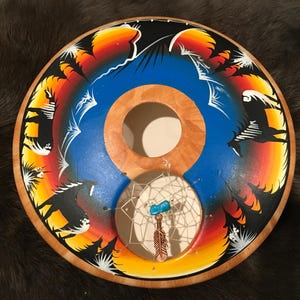 Pode incluir: Um apanhador de sonhos redondo de madeira com um design pintado em azul, amarelo e laranja. O apanhador de sonhos tem uma pequena teia de contas brancas com uma pedra turquesa e uma pena pendurada.
