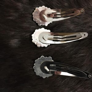 May include: Three silver-toned metal hair clips, each featuring a circular, scalloped-edge decorative piece attached to a standard snap-style barrette mechanism. The clips are arranged vertically, showing the underside of the metal hardware.