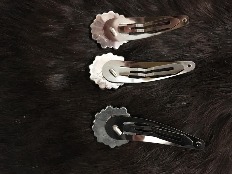 May include: Three silver-toned metal hair clips, each featuring a circular, scalloped-edge decorative piece attached to a standard snap-style barrette mechanism. The clips are arranged vertically, showing the underside of the metal hardware.