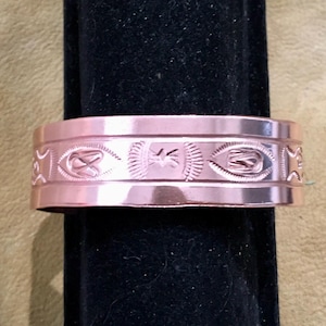 May include: A copper-colored cuff bracelet with an engraved design. The bracelet features repeating patterns of stylized shapes and symbols. The metal has a polished finish, reflecting light. The bracelet is displayed on a black surface.