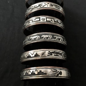 May include: A collection of silver-toned metal bracelets with intricate etched designs. The bracelets feature various patterns, including geometric shapes, animal figures, and stylized landscapes. Each bracelet has a dark, oxidized background, enhancing the details.