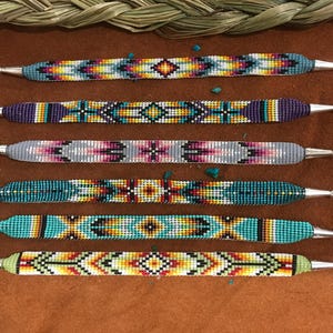 May include: Six beaded bracelets with silver-toned clasps. The bracelets feature intricate geometric patterns in various colors, including turquoise, yellow, red, and purple. The bracelets are displayed on a brown surface.
