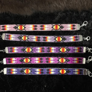Könnte beinhalten: Fünf Armbänder mit Perlen in Rosa-, Lila- und Blautönen. Jedes Armband hat ein sich wiederholendes Muster aus orangefarbenen, gelben und schwarzen Perlen. Die Armbänder haben silberfarbene Verschlüsse und Kettenverlängerungen.