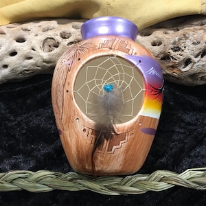 Puede incluir: Un jarrón de cerámica marrón hecho a mano con un diseño de atrapasueños. El jarrón presenta una abertura redonda con una red tejida, una cuenta azul y una pluma. El jarrón tiene un borde púrpura y está decorado con patrones de inspiración nativa americana y una escena de puesta de sol.