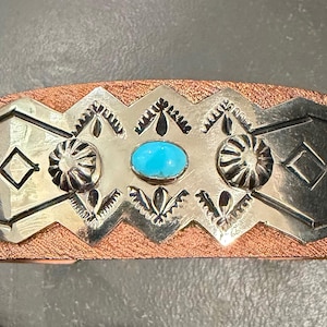 May include: A cuff bracelet featuring a textured copper-colored band overlaid with a polished silver-tone metal plate. The plate displays engraved geometric patterns, two circular embossed accents, and a central oval turquoise stone.